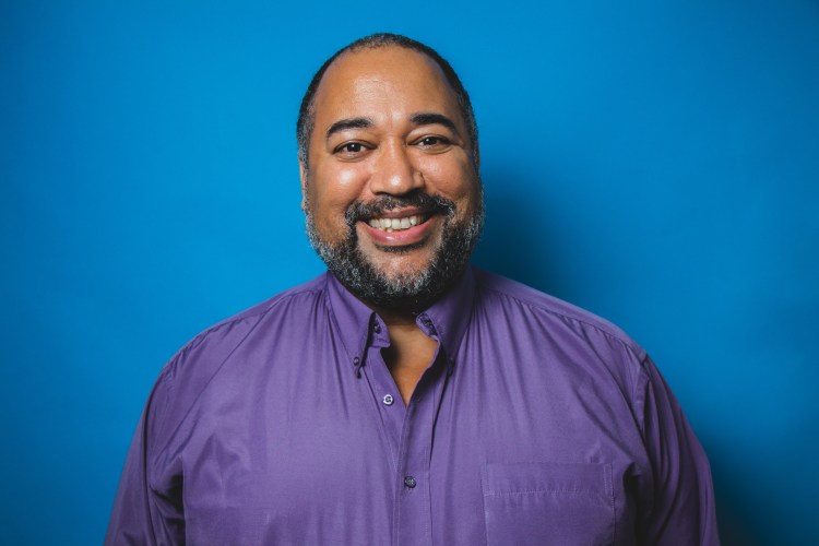 Smiling profile image of Velvet Wells as The Velvet Duke on a gradient blue background. In the image, Velvet is a Black man smiling to the camera and wearing a purple dress shirt. Photo: Tyra Sweet (2019)