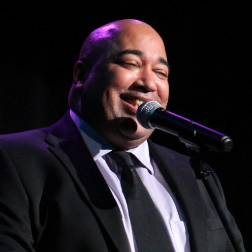 Velvet Wells Mirth photo: Krissia Valiente An image of Velvet Wells, a Black man in a black suit, smiling into a microphone. Black curtains are behind him, a purple light shines on his head and shoulders.