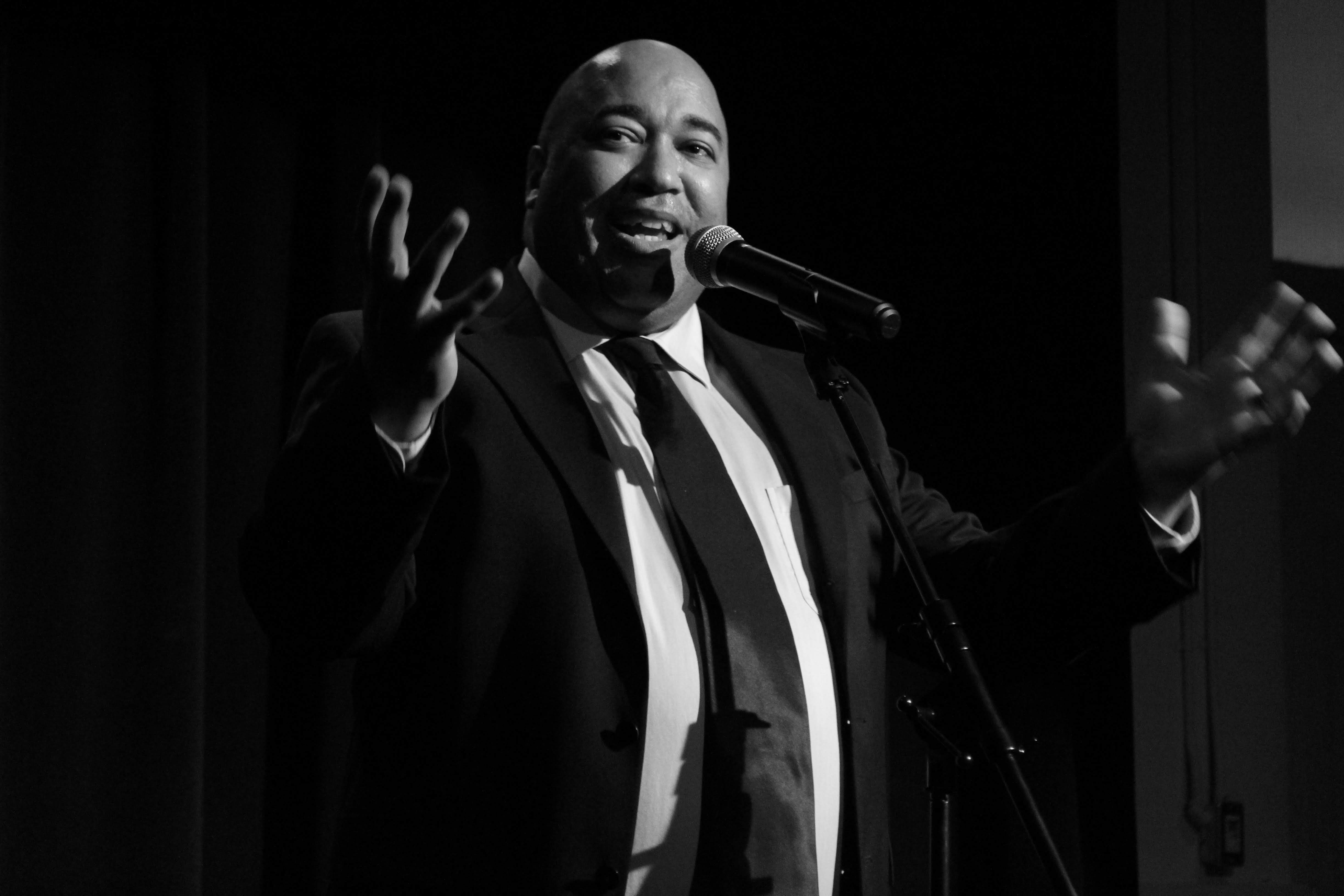 Grayscale image of Velvet Wells, a Black male in a black suit, speaking, into a microphone with a smile on his face. photo: Krissia Valiente