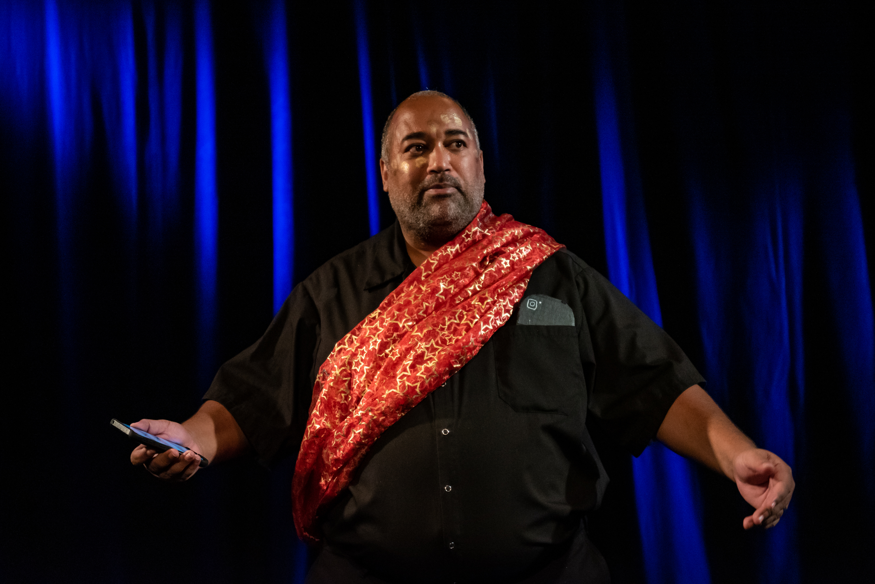 Velvet shares a Land Acknowledgement and call to action with the audience. They are a Black fat person with salt and pepper hair. They are wearing black clothing and a red sash across their body. Behind them, electric blue lights diagonally cascade across black curtains. photo: Stoo Metz Photography