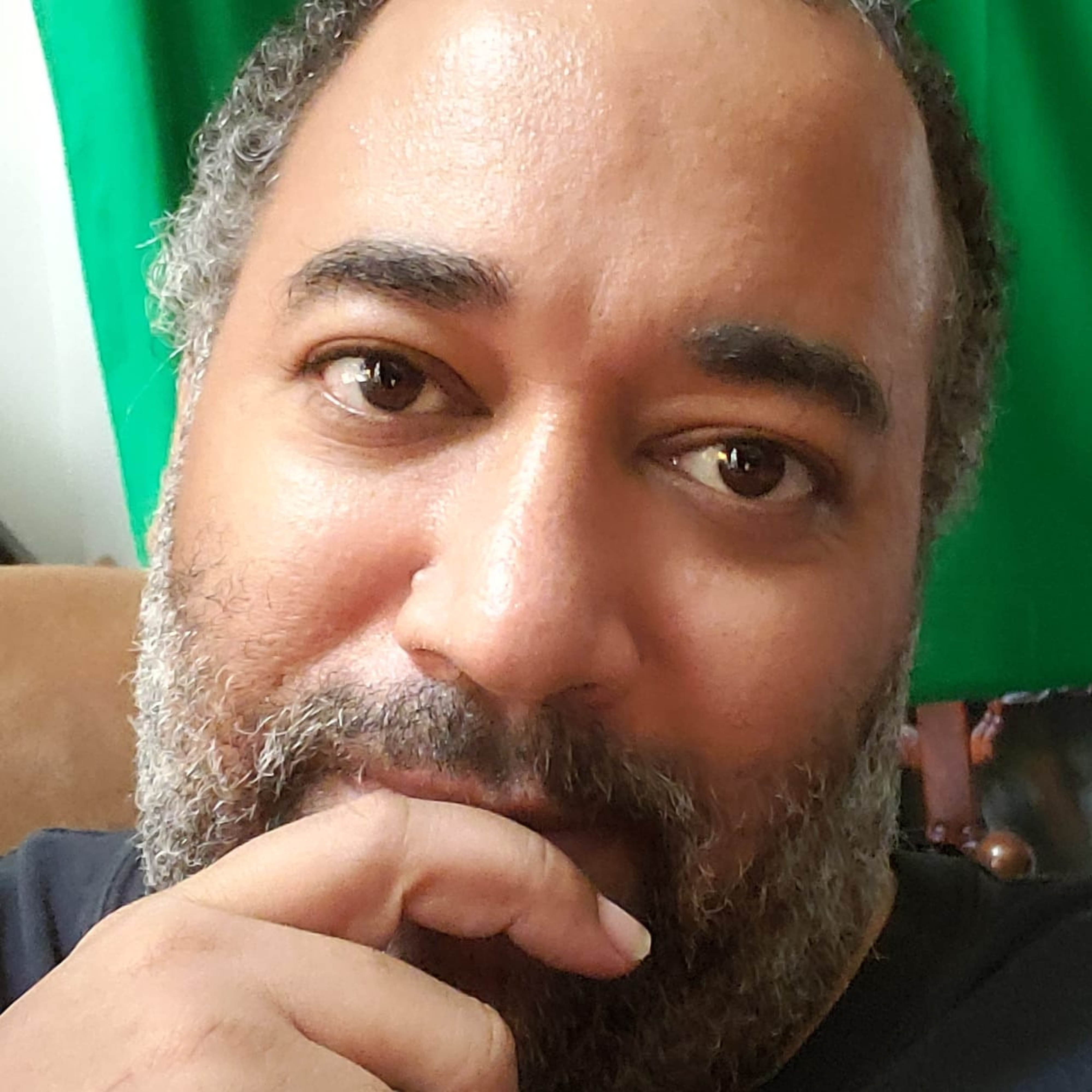 Velvet is a Black fat person with salt and pepper hair and beard. Their index finger rests on their lips in contemplation. The look in their brown eyes is warm and inviting. Behind Velvet is a haphazard green screen. photo: Velvet