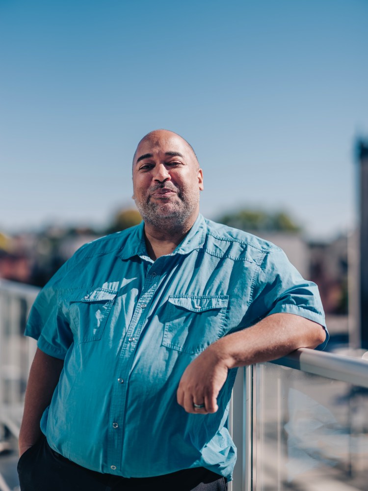 Velvet Wells (Ottawa) stands on the balcony of the Ottawa Art Gallery, the city and skyline blurred behind them. Captured by Curtis Perry Photography (2022)