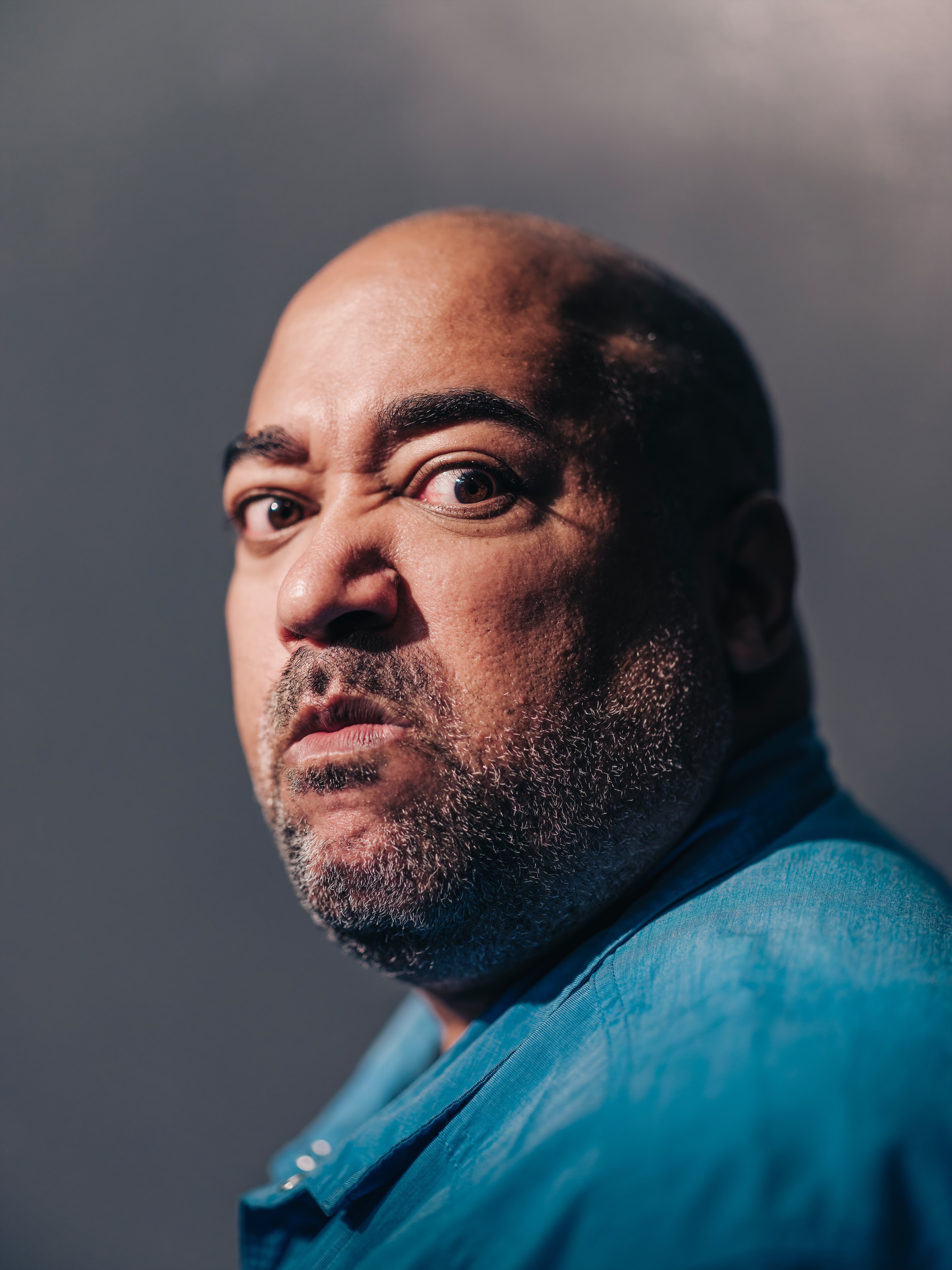 Velvet looks directly at you, feeling very upset. This is acting -- they are happy you're seeing this image. They are wearing a teal shirt to accentuate their Black skin and the blurred gray background. Captured by Curtis Perry (2022)