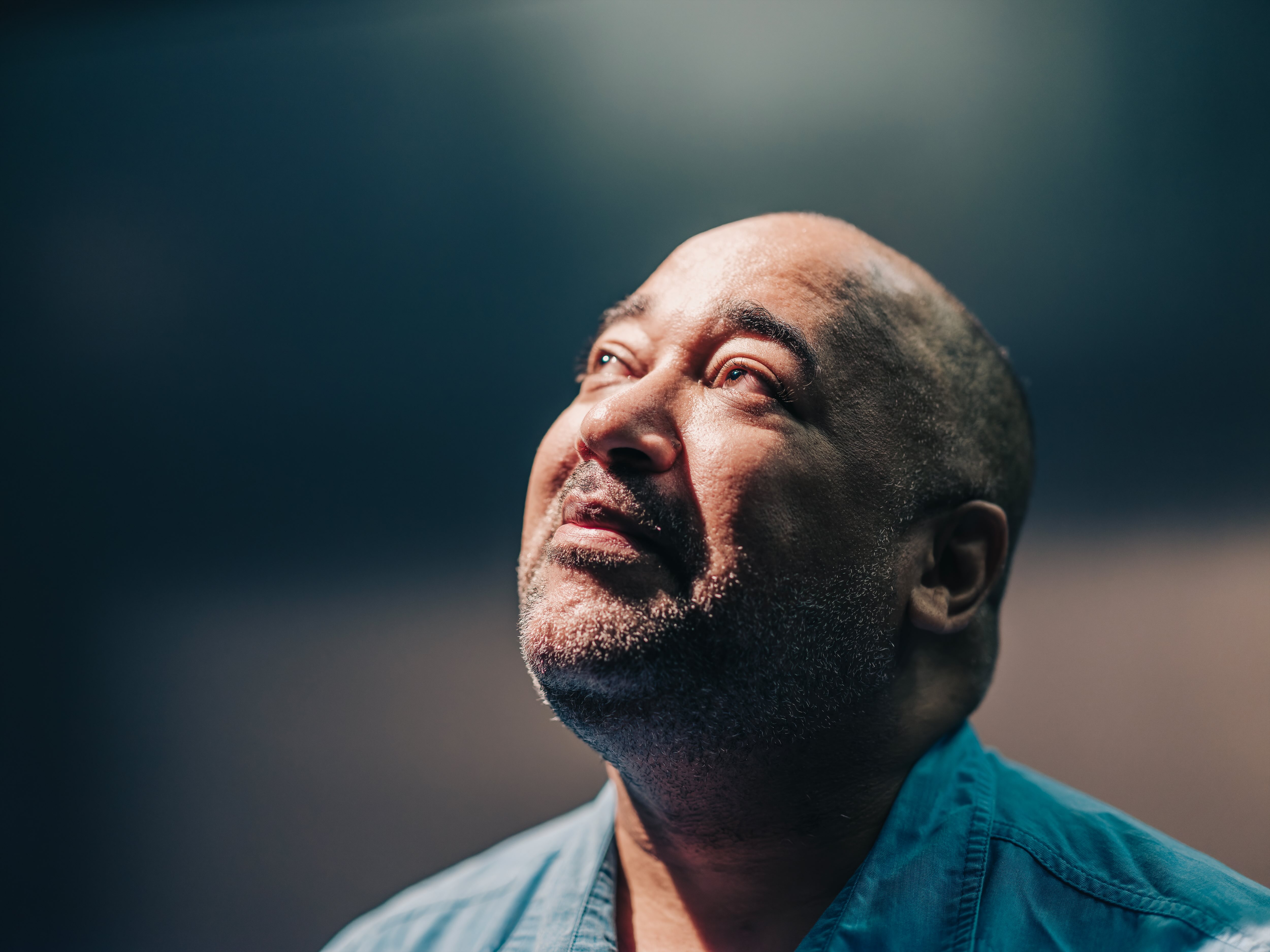Velvet looks wistfully upward toward the top-left; shadows glance their left side, their right eye fully in the light. Their teal shirt and the blurred gray background enhance the focus on Velvet's expression. Captured by Curtis Perry (2022)