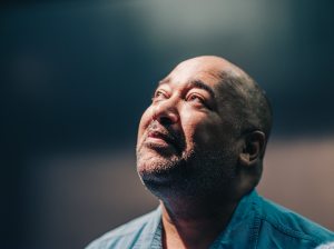 Velvet looks wistfully upward toward the top-left; shadows glance their left side, their right eye fully in the light. Their teal shirt and the blurred gray background enhance the focus on Velvet's expression. Captured by Curtis Perry (2022)