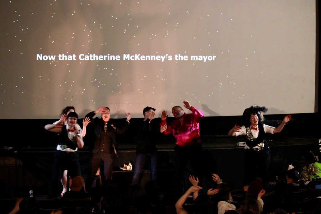 ID: Catherine McKenney and The Velvet Duke smile together mid-song; Velvet has a raspberry shimmery shirt which stands out amongst the traditional black and white style of NYE. "Now that Catherine McKenney's the mayor" is on the screen behind the singing ensemble Small Fish Show (IG: smallfishshow) PHoto: Theresa Graham Studio (IG: theresagrahamstudio)