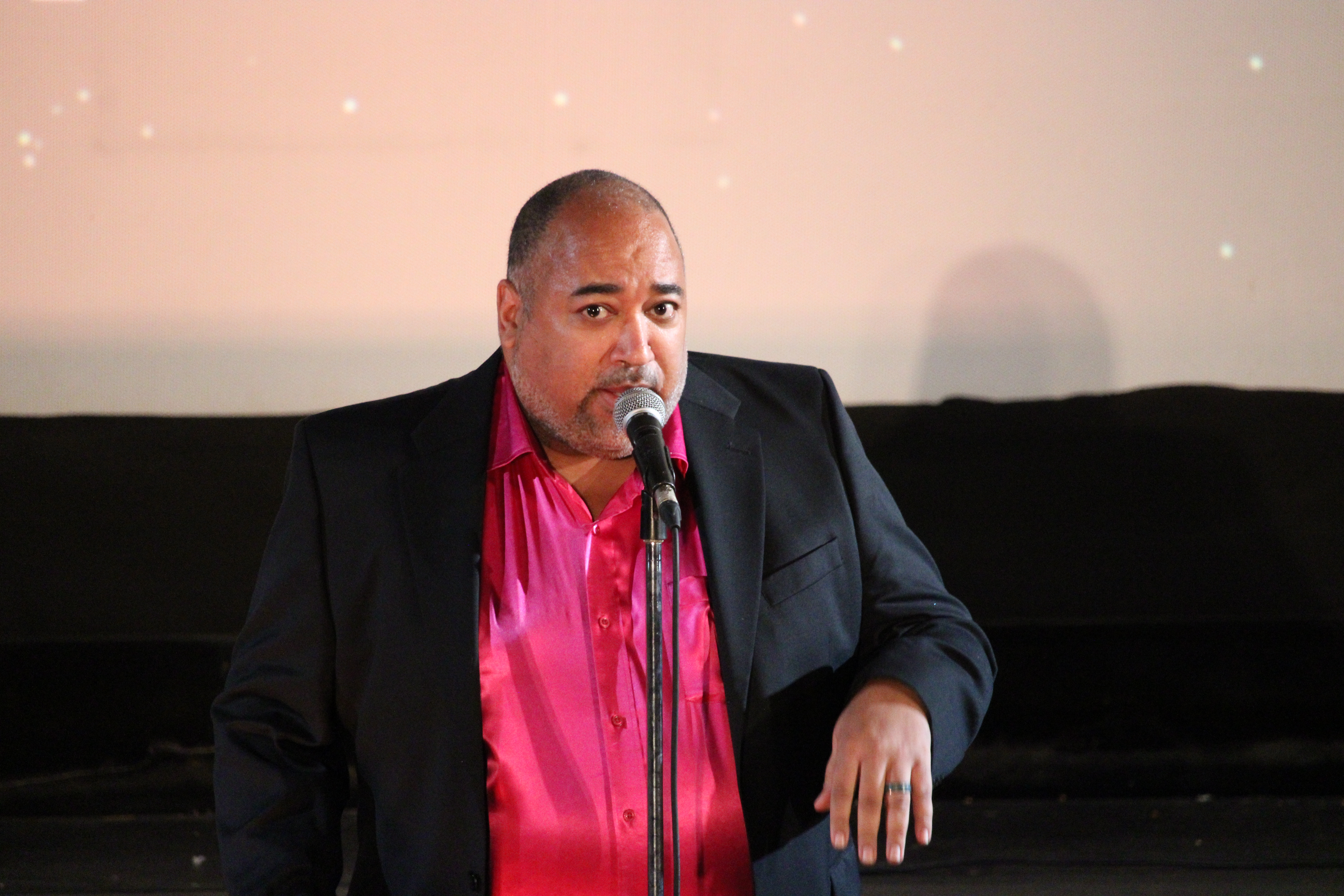 ID: The Velvet Duke, wearing a raspberry shimmery shirt underneath a black jacket, looks up at the audience in one of the few times they aren't smiling Small Fish Show (IG: smallfishshow) PHoto: Theresa Graham Studio (IG: theresagrahamstudio)