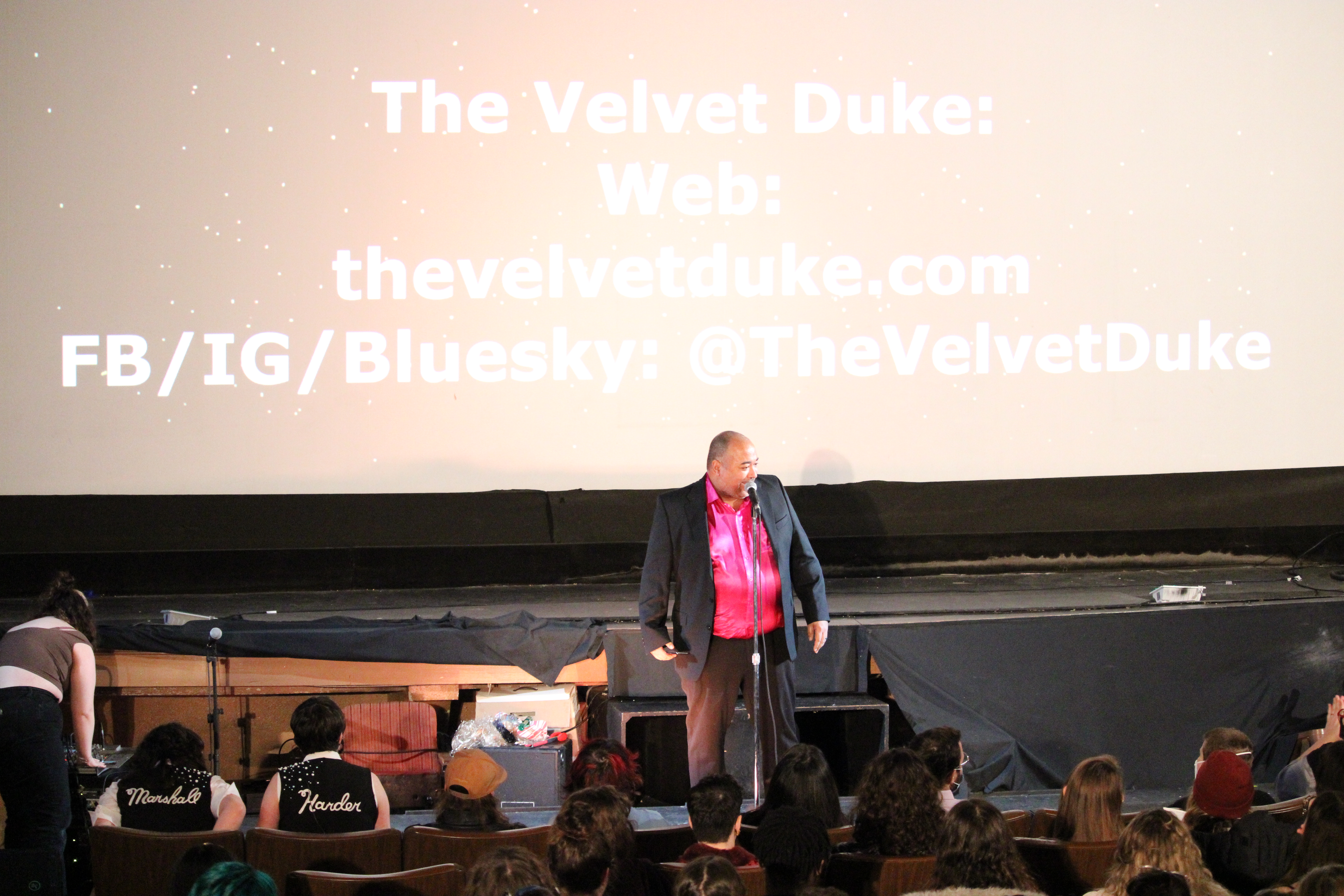 ID: The screen behind The Velvet Duke has their social media contact information. They are wearing a raspberry shimmery shirt underneath a black jacket, responding to one of the front row audience members Small Fish Show (IG: smallfishshow) PHoto: Theresa Graham Studio (IG: theresagrahamstudio)