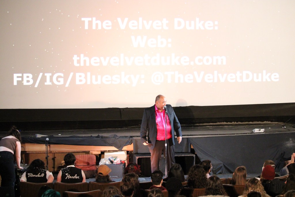 ID: The screen behind The Velvet Duke has their social media contact information. They are wearing a raspberry shimmery shirt underneath a black jacket, responding to one of the front row audience members
Small Fish Show (IG: smallfishshow) Photo: Theresa Graham Studio (IG: theresagrahamstudio)