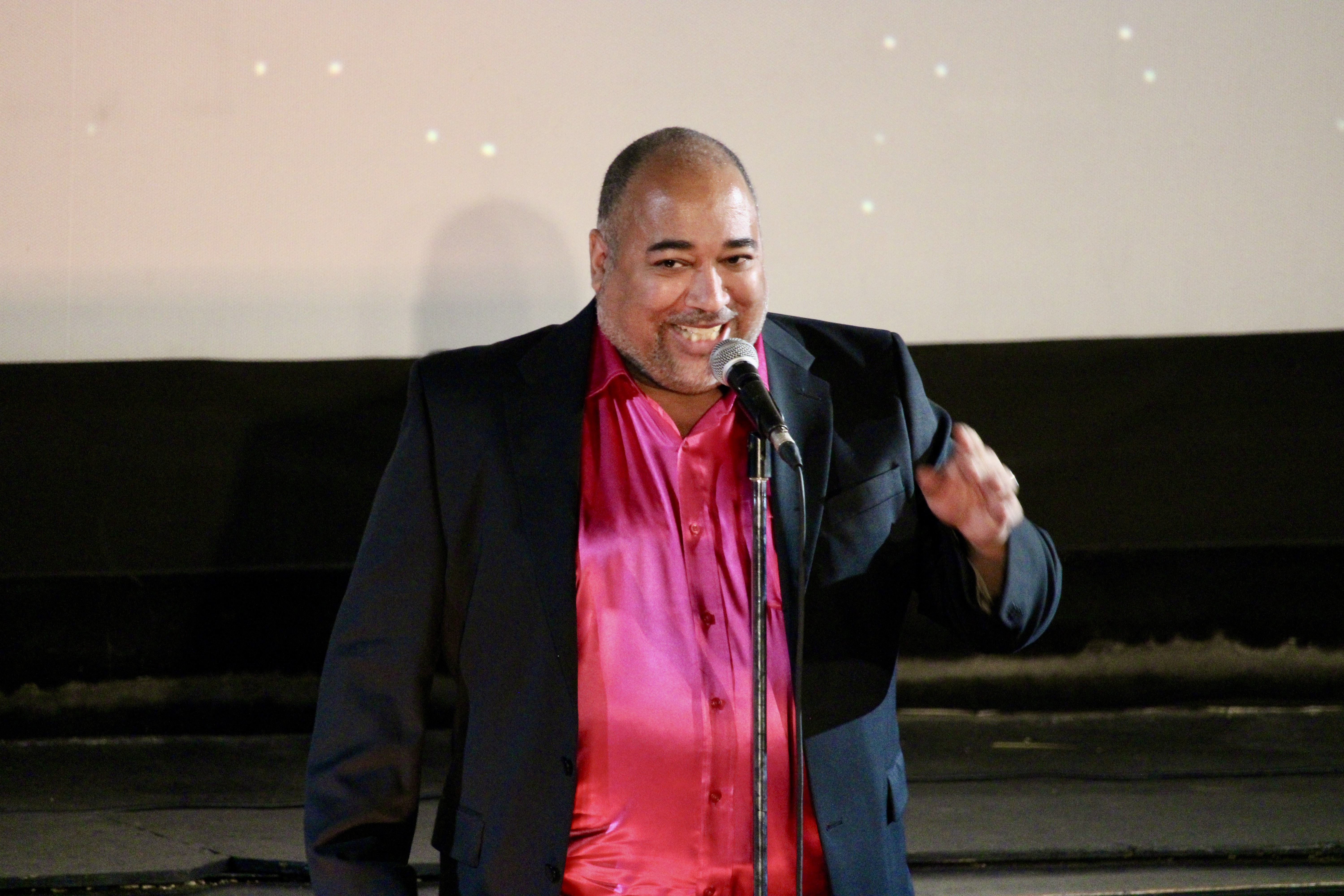 ID: The Velvet Duke is wearing a raspberry shimmery shirt underneath a black jacket, and laughing with the audience Small Fish Show (IG: smallfishshow) PHoto: Theresa Graham Studio (IG: theresagrahamstudio)