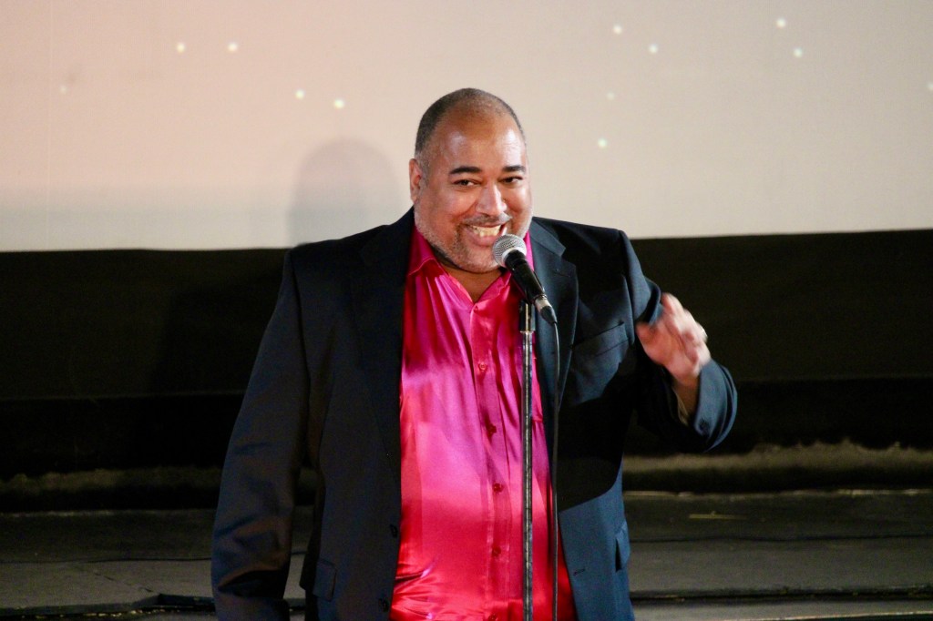 ID: The Velvet Duke is wearing a raspberry shimmery shirt underneath a black jacket, and laughing with the audience 
Small Fish Show (IG: smallfishshow) Photo: Theresa Graham Studio (IG: theresagrahamstudio)