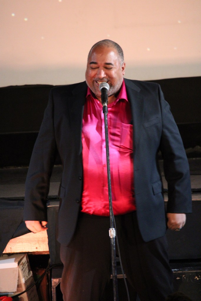 ID: The Velvet Duke is wearing a raspberry shimmery shirt underneath a black jacket, laughing with eyes closed 
Small Fish Show (IG: smallfishshow) Photo: Theresa Graham Studio (IG: theresagrahamstudio)