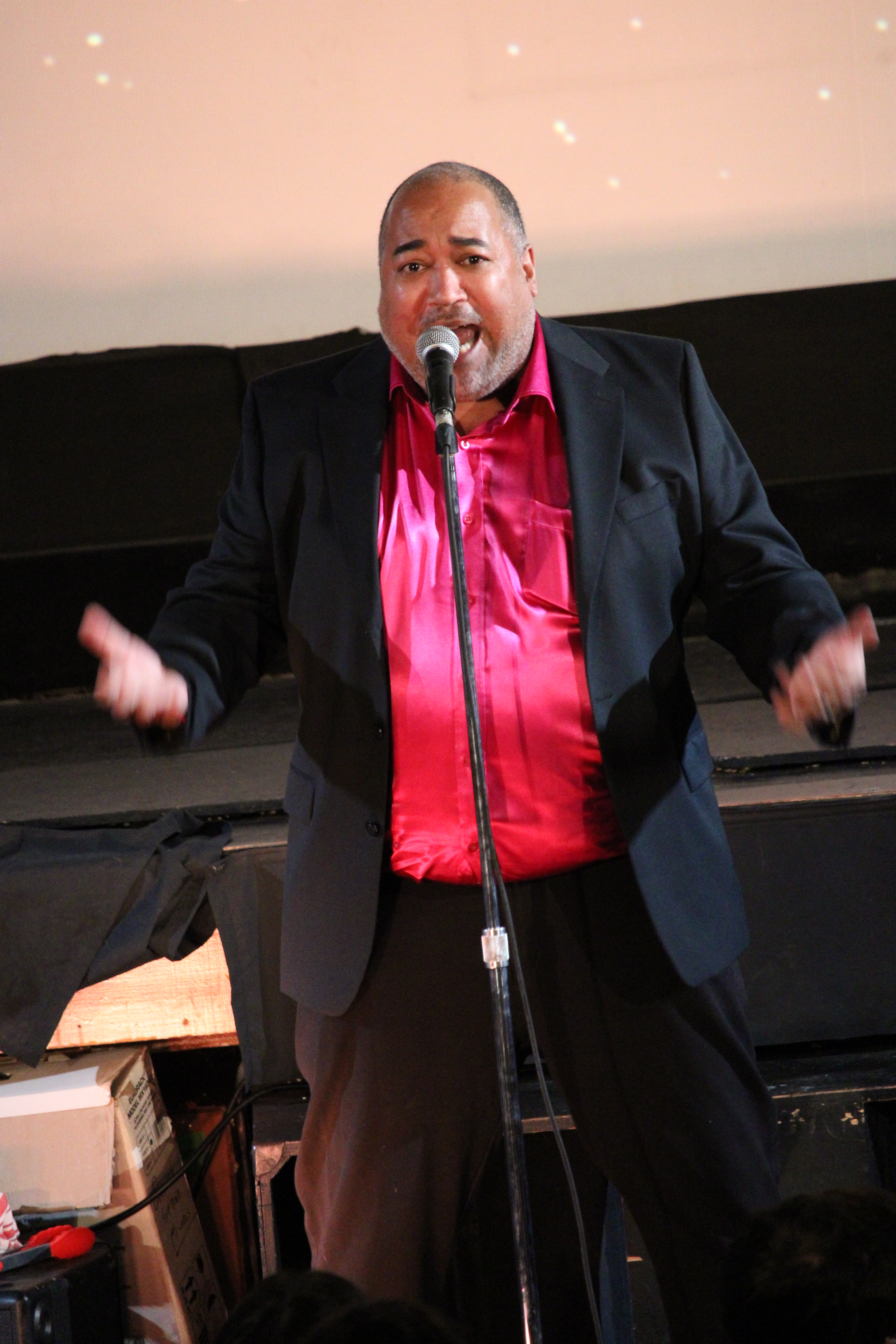 ID: The Velvet Duke is wearing a raspberry shimmery shirt underneath a black jacket, sings with arms outstretched to the audience Small Fish Show (IG: smallfishshow) PHoto: Theresa Graham Studio (IG: theresagrahamstudio)