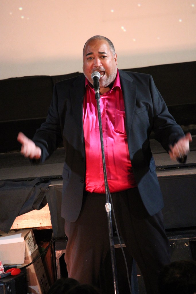 ID: The Velvet Duke is wearing a raspberry shimmery shirt underneath a black jacket, sings with arms outstretched to the audience 
Small Fish Show (IG: smallfishshow) Photo: Theresa Graham Studio (IG: theresagrahamstudio)