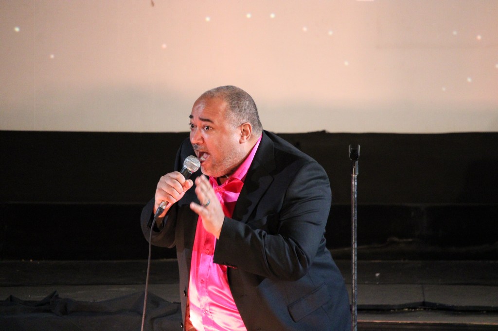 ID: The Velvet Duke is wearing a raspberry shimmery shirt underneath a black jacket, singing and leaning toward the audience 
Small Fish Show (IG: smallfishshow) Photo: Theresa Graham Studio (IG: theresagrahamstudio)