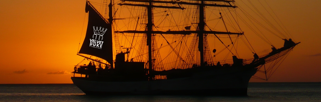 A schooner sails with the orange sunset behind. The Velvet Duke Productions logo flies on the rear sail